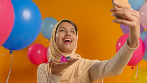 Retrato-De-Estudio-De-Una-Mujer-Tomándose-Un-Selfie-Usando-Hijab-Celebrando-Un-Cumpleaños-Soplando-Confeti-De-Papel-Rodeado-De-Globos